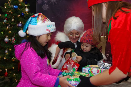 Young girl and boy receiving gifts from an elf while sitting with Mrs. Claus